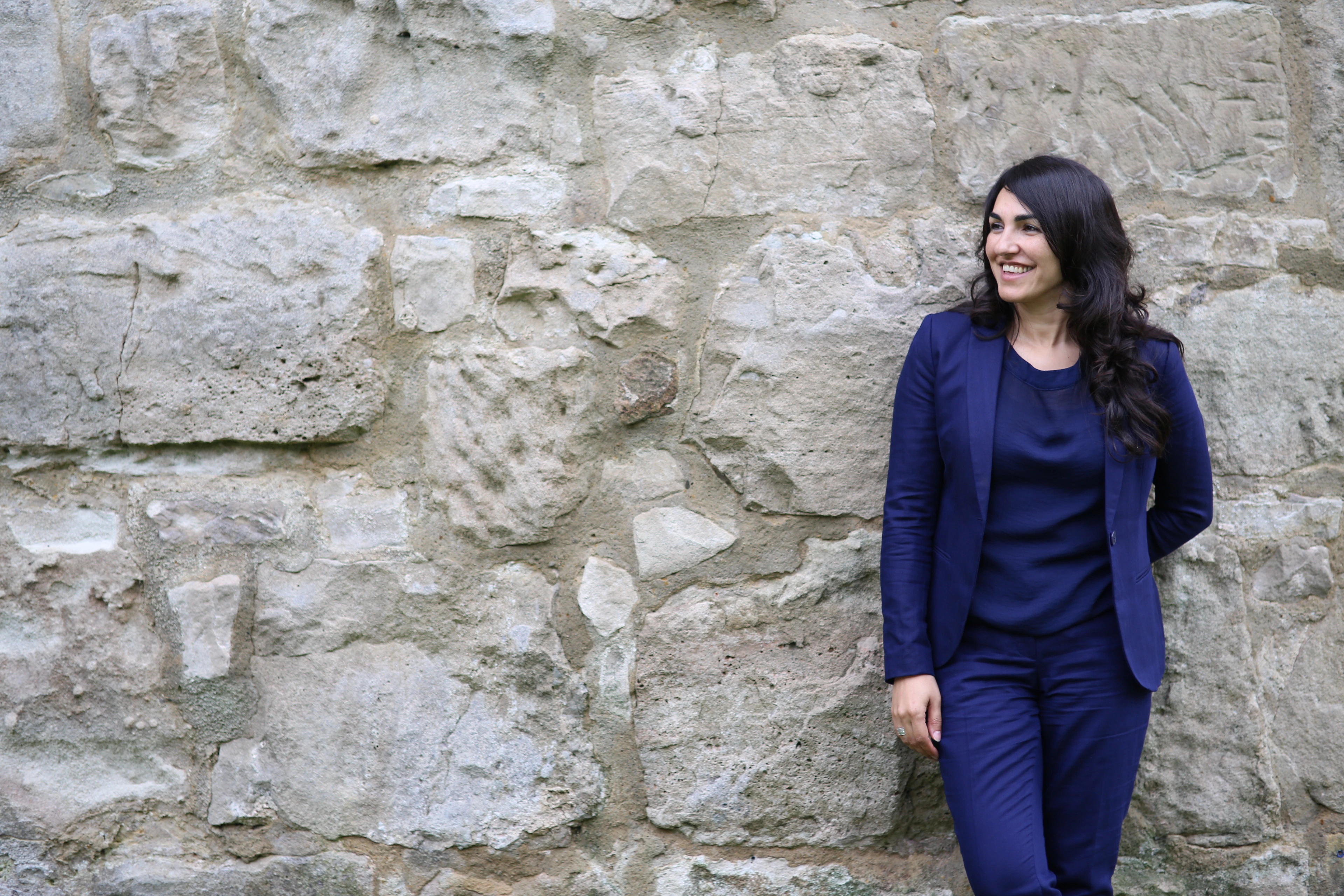 Asiye Balıkçı-Schmidt lehnt an einer hellen Natursteinmauer und blickt zur Seite