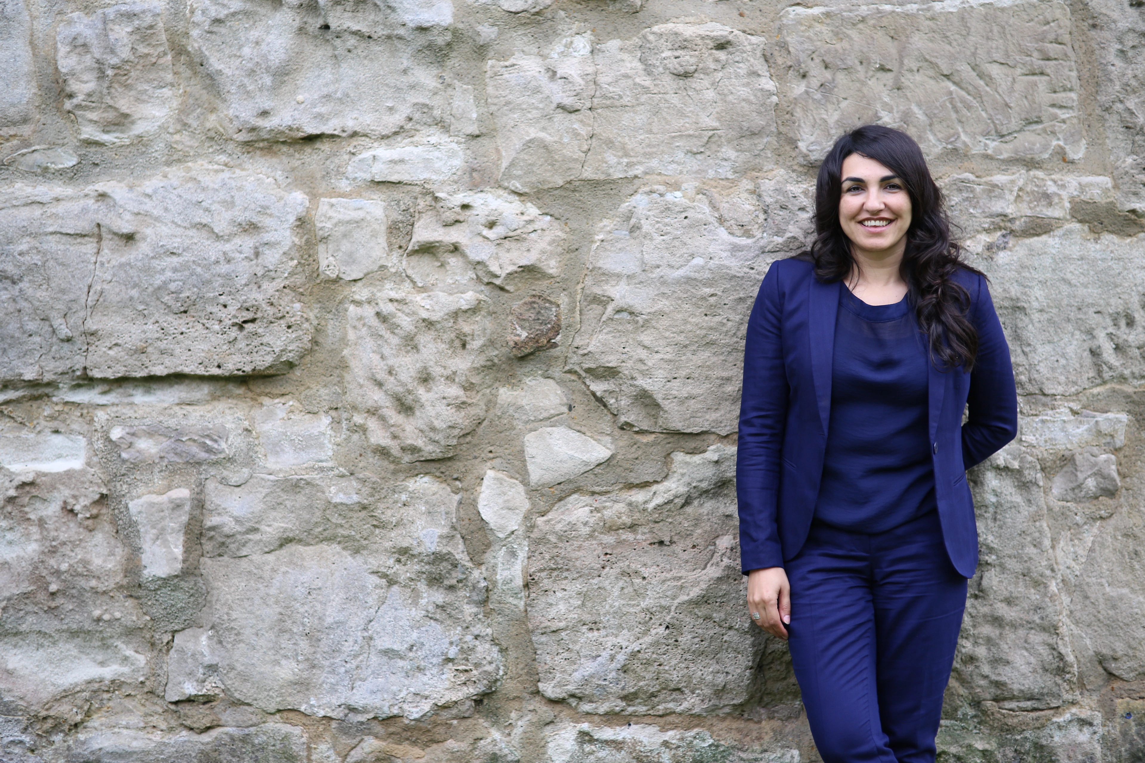 Asiye Balıkçı-Schmidt vor einer großen Natursteinmauer, den Blick nach vorn gerichtet