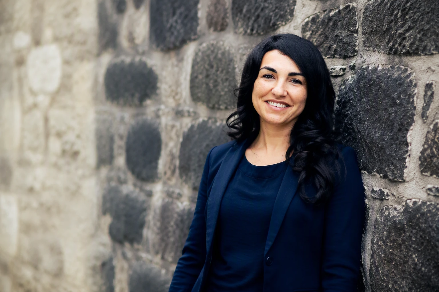 Asiye Balıkçı-Schmidt, lächelnd an einer hellen Natursteinmauer